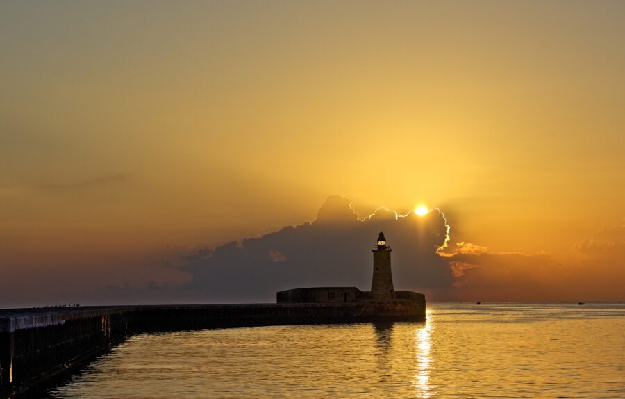 Stunning Malta sunset over the sea, with a silhouette of a lighthouse and harbour wall. Golden light and vibrant colours paint the sky.