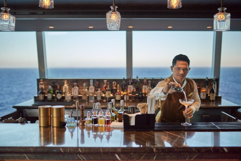 A skilled bartender crafts an exquisite cocktail at a stylish bar, overlooking the stunning ocean view. Travel Escapes