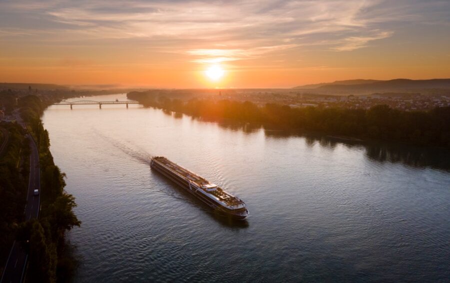 A breathtaking sunset view over the Danube River, with a river cruise ship gently sailing along. The warm hues of orange and pink paint the sky and reflect on the water. Experience the beauty of a Balkan river cruise.