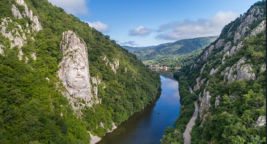 Majestic view of the Danube River flowing through the dramatic Iron Gates Gorge, with towering cliffs and lush greenery lining the banks under a bright sky. Experience the natural wonders of the Balkans on a river cruise.