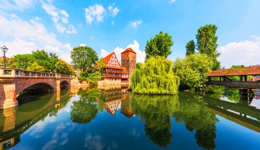 Picturesque scene in Nuremberg, Germany, featuring charming half-timbered buildings and a historic tower reflected in the calm waters of the Pegnitz River under a partly cloudy blue sky. Experience the beauty of Bavaria on your European river cruise.