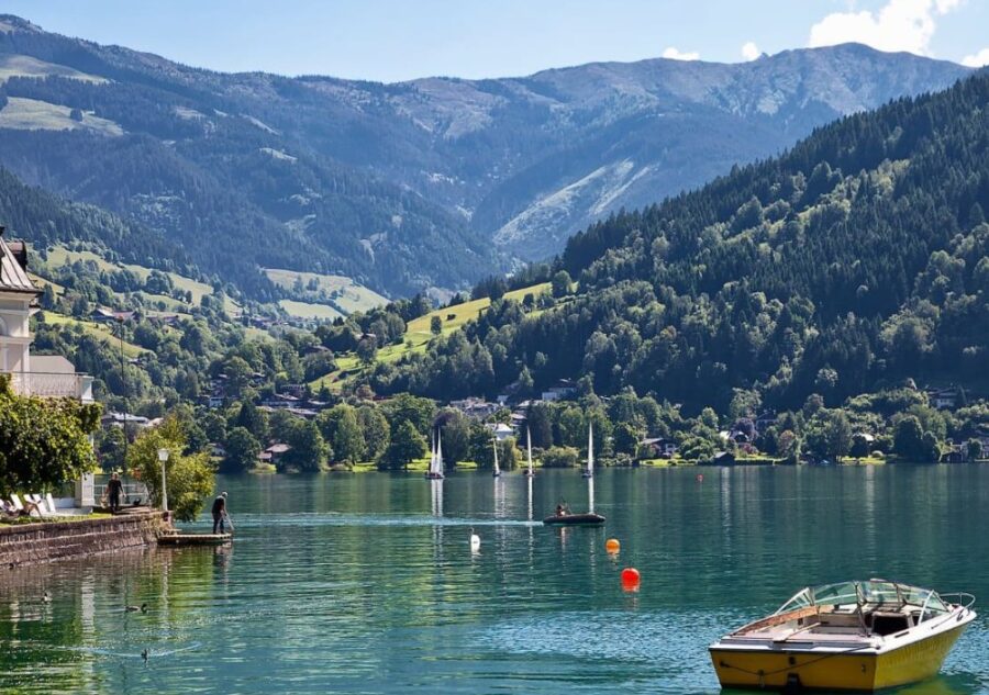 A picturesque lake in the Austrian Alps with green mountains, sailing boats on the water, and a person on a jetty, under a blue sky.