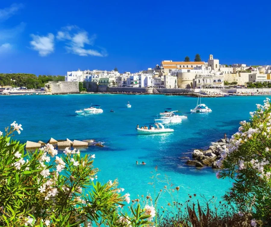 A scenic view of Otranto, Puglia, showing the whitewashed town, ancient walls, and boats floating on vibrant turquoise water in the bay.