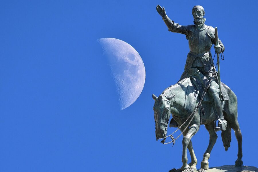 A bronze equestrian statue of a knight raising his hand, set against a clear blue sky with a large half-moon visible in the background.