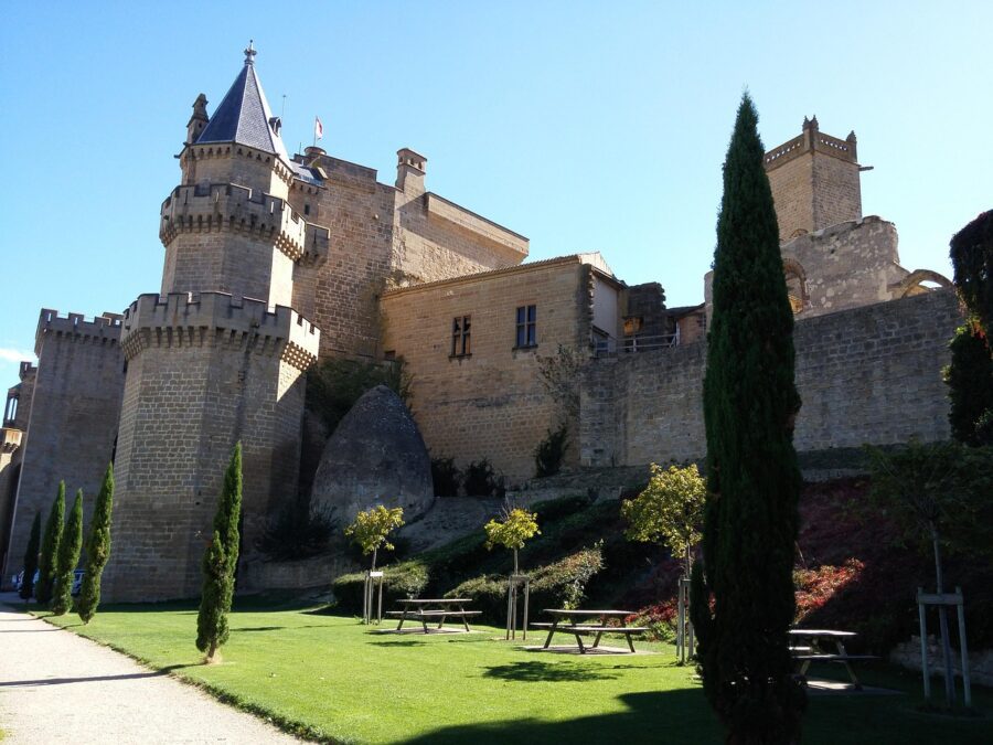 The towering stone walls and turrets of the Royal Palace of Olite under a bright blue sky, with manicured green lawns and trees in the foreground.